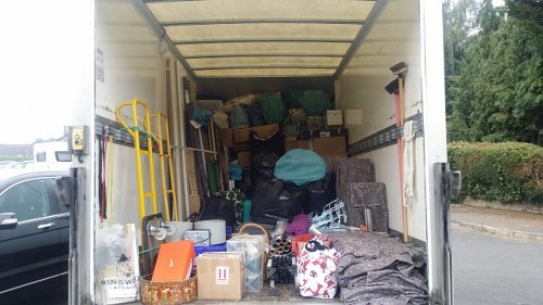 The image shows the interior of a rear cargo van parked on a driveway or street, filled with a variety of discarded household items and rubbish. In the foreground, there are several cardboard boxes, some filled with miscellaneous objects, and a small ornamental lantern. To the right, a rolled-up carpet or mat with a dark, textured surface is visible. Towards the back, numerous large black and dark green refuse bags are stacked, along with cardboard boxes and plastic containers, some wrapped in or covered by light blue and teal fabric or plastic. On the left side, a yellow trolley or hand truck leans against the van's wall, and a small orange plastic container is seen placed among the rubbish. The van's interior ceiling is light-colored, and the side walls are metallic. Outside, the background shows greenery and part of a paved driveway or street, indicating the scene is located in a residential or semi-urban area, likely serviced by Commercial Waste Battersea for rubbish removal services in the Battersea or larger London area. The space appears to be organized in preparation for waste disposal or collection.