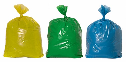 Three large, tightly knotted plastic rubbish bags are positioned in a row on a paved outdoor surface, with a yellow bag on the left, a green bag in the middle, and a blue bag on the right. The bags are made of glossy, flexible plastic, with slight creases and wrinkles visible on their surfaces, and each has the top gathered and tied off with a visible knot. The background shows a section of concrete paving or driveway, typical of an outdoor area in Battersea, London, suitable for rubbish collection. The bags appear full and weighty, indicating they contain household or commercial waste ready for removal. The scene is evenly lit, with natural daylight accentuating the bright, vibrant colours of the plastic bags, and the environment is clean and unobstructed, representing an active rubbish disposal setting managed by Commercial Waste Battersea.