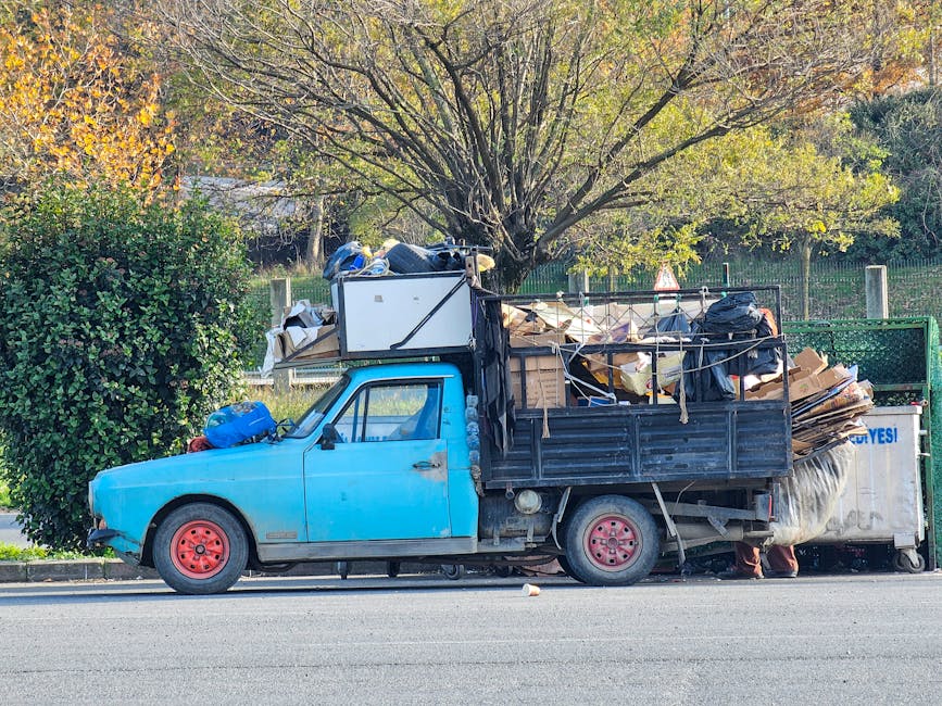 A small, blue, vintage-style flatbed truck parked along the side of a street with a large load of mixed household rubbish and debris. The truck's body is painted in a faded blue color, with exposed re