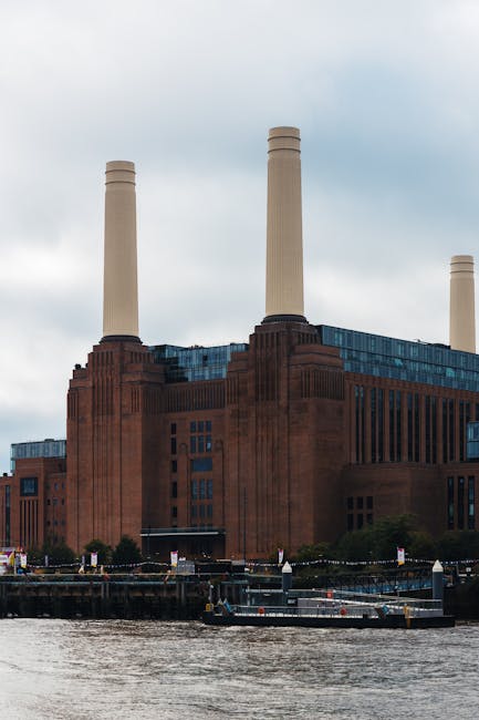 The image depicts a riverside scene with a large industrial-style building in the background, featuring multiple tall chimneys and brickwork textures, set against a cloudy sky. In the foreground, part