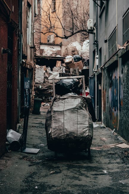 A row of large, wheeled rubbish bins arranged along a pavement beside a brick wall, with their lids closed. The bins are primarily blue, with the green bin closest to the viewer, and have matching col