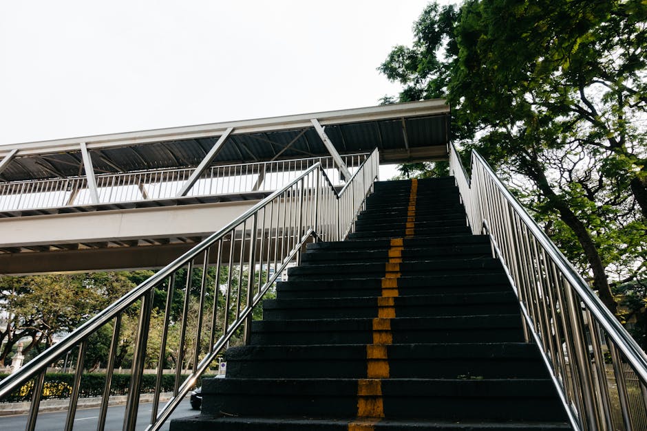 A weathered concrete staircase with uneven, stained steps leads upward, flanked on the left by a yellow metal handrail showing chipped paint and rust patches. Adjacent to the base of the stairs, a lar
